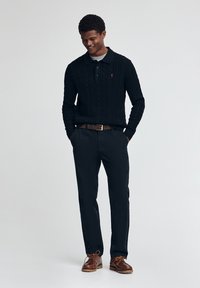 Svart kabelstickad tröja med krage, i kombination med svarta byxor och bruna snörskor. Noterbar röd logotyp på tröjan.