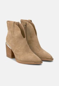 Botas de tobillo de ante en beige con puntera afilada, cierre de cremallera y un tacón cuadrado que presenta un acento metálico en el lateral.