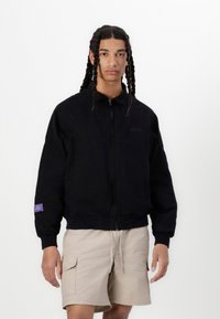 Svart zip-up jakke med ribbet mansjetter og kant, med en liten logo på brystet og et lilla merkelapp på ermet. Brukt over beige shorts.