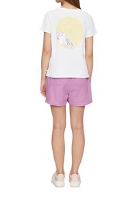 Witte katoenen t-shirt met gele zon grafiek en tekst op de achterkant, gecombineerd met lichtpaarse linnen shorts. Casual, comfortabele outfit.