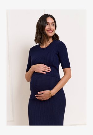 Terhes nő egy tengerészkék bordázott ruhában óvatosan tartja a hasát, mosolyogva, oldalra nézve, egyszerű, világos háttér előtt.