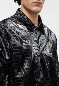 Čierna lesklá vetrovka s geometrickými vzormi. Má golier, zapínanie na gombíky a zlaté logo akcenty. Materiál má lesklú textúru.