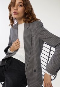 Szary żakiet w paski z klapami i zapięciem na guzik, noszony na białej koszuli i czarnych spodniach, z uwydatnionym dopasowaniem i zorganizowanym krojem.