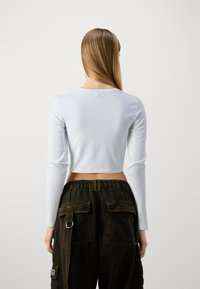 Kvinde med langt, glat hår iført en hvid langærmet cropped trøje og løse mørke cargo bukser, stående mod en ensfarvet lys baggrund, med ryggen vendt mod kameraet.