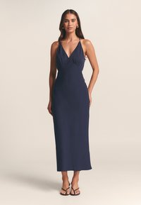 Tamsiai mėlyna, be rankovių maksi suknelė su gilia V formos iškirpte ir apvyniojamu bodžiu, pagaminta iš lygaus audinio, pasižymi tiesia silueto linija ir kulkšnies ilgiu.
