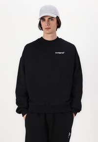 Sort sweatshirt med ribbede manchetter og kant, der har et hvidt logo på brystet. Parret med en lys grå kasket og mørke bukser.