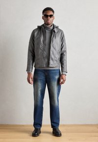 Graue Lederjacke mit Kapuze, grauer Rollkragenpullover und blaue Jeans, kombiniert mit schwarzen Schuhen und Sonnenbrille. Schlichtes, gerades Design.