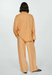 Persona con capelli scuri lunghi fino alle spalle, indossante una camicia a maniche lunghe di un marrone chiaro e ampipantaloni abbinati, in piedi di spalle.
