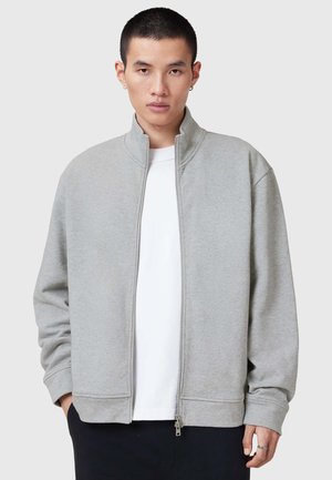 Ljusgrå zip-up sweatshirt med hög krage, ribbat manschetter och avslappnad passform, lager på en vit T-shirt. Slät tygtextur.
