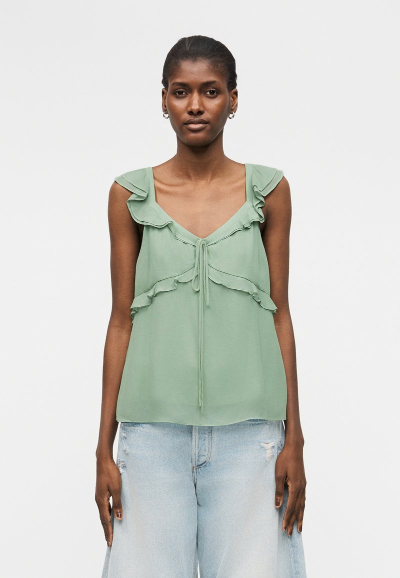 Γυναίκα που φοράει ένα αμάνικο τοπ σε χρώμα φασκόμηλου με βολάν και δεσίματα μπροστά, συνδυασμένο με ανοιχτό μπλε σκισμένο παντελόνι φαρδιάς γραμμής.