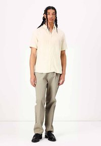 Korte mouwen, lichtbeige shirt met een gestructureerd oppervlak, gecombineerd met lichtgroene gestructureerde broek. Model draagt zwarte schoenen.
