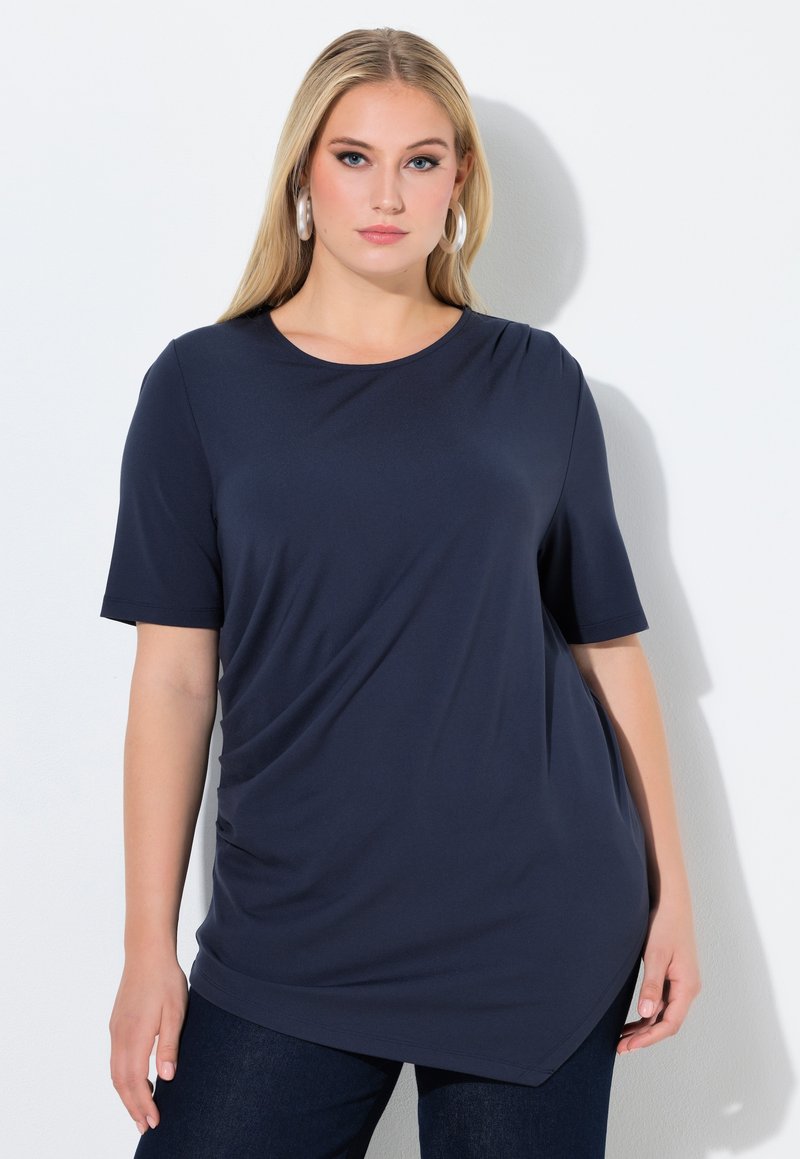 Femme blonde avec des boucles d'oreilles créoles portant un haut bleu marine à manches courtes avec un ourlet asymétrique et un jean foncé, debout contre un mur blanc.