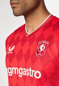 Červený futbalový dres s diamantovým vzorom, krátkymi rukávmi, bielym lemovaním a výstrihom do V. Obsahuje logo F.C. Twente a text "gmgastro".