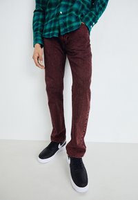Persona vestida con una camisa de cuadros verdes, jeans rojos oscuros y zapatillas negras con suelas blancas, de pie contra un fondo blanco liso.