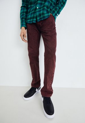 Persona vestida con una camisa de cuadros verdes, jeans rojos oscuros y zapatillas negras con suelas blancas, de pie contra un fondo blanco liso.
