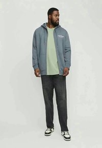 Homem com um hoodie azul com fecho, t-shirt verde clara, jeans escuros e sapatilhas preto e branco, em pé e olhando para o lado.