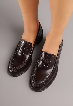 Mocassins en cuir brun foncé brillant avec des clous en métal, portés pieds nus sur un fond gris uni.