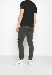 Czarny t-shirt z krótkim rękawem, oliwkowe spodnie typu cargo z wieloma kieszeniami, białe trampki z czerwonymi akcentami, jednolite tło.