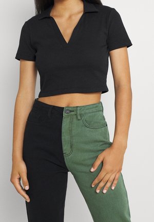 Vrouw die een zwarte korte mouw cropped polo en twee-kleurige broek draagt met één groene en één zwarte broekspijp, staand met één hand op haar heup.