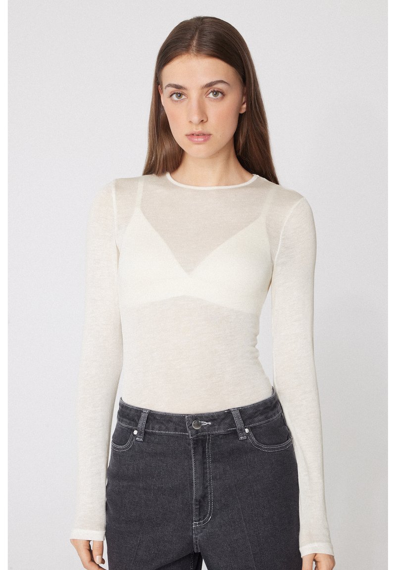 Junge Frau mit langen braunen Haaren, die ein durchsichtiges weißes Langarm-Top über einem weißen BH und dunkelgrauen hoch taillierten Jeans trägt.