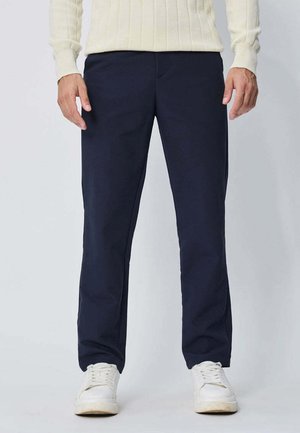 Homme portant un pantalon bleu marine, des baskets blanches et un pull côtelé couleur crème, debout devant un fond clair uni.