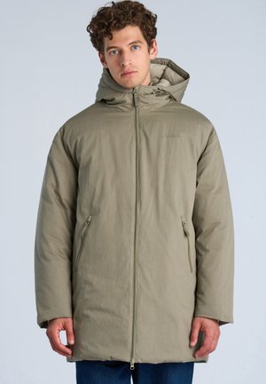 Jonge man met krullend haar, die een beige winterjas met capuchon draagt met een rits aan de voorkant en ritssluitingen op de zakken, tegen een egale lichte achtergrond.