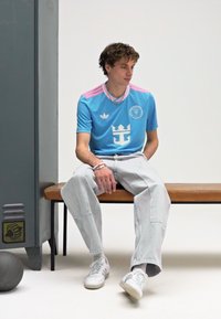 Camisola azul-clara de manga curta com detalhes em rosa e logótipo de coroa, combinada com jeans cinzentos claros e largos e ténis brancos, sentado num banco.