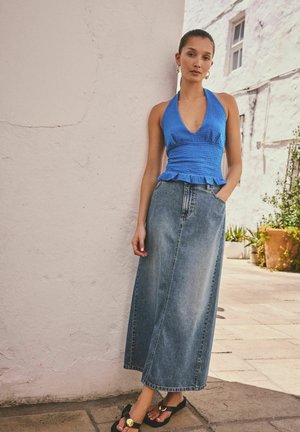 Sieviete zilā haltera tipa topā, garā džinsa svārkā un melnās sandales, stāvot pie baltas sienas ārpus telpām ar tuvumā esošiem augiem.