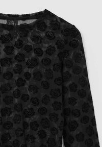 Haut en maille noire avec un motif de roses en relief, manches longues, encolure ronde et texture transparente. Un éclat subtil ajoute de la profondeur au design floral.