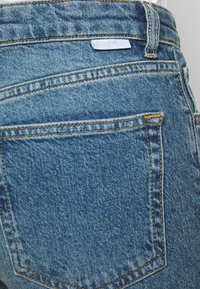 Jean en denim bleu délavé avec une surface texturée. Comprend deux poches arrière avec des coutures jaunes et une petite étiquette portant la mention "Boyish."