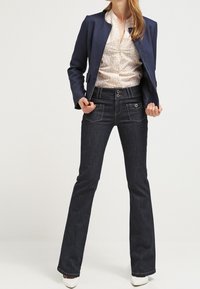 Blazer blu navy, blusa a fantasia, jeans flare in denim scuro, bottoni lucenti, vestibilità elegante e scarpe bianche; design minimalista con linee pulite.