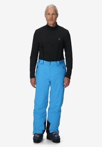 Man in zwarte lange mouwen shirt en felblauwe skibroek met bretels en skischoenen, rechtop staand met het gezicht naar voren op een witte achtergrond.