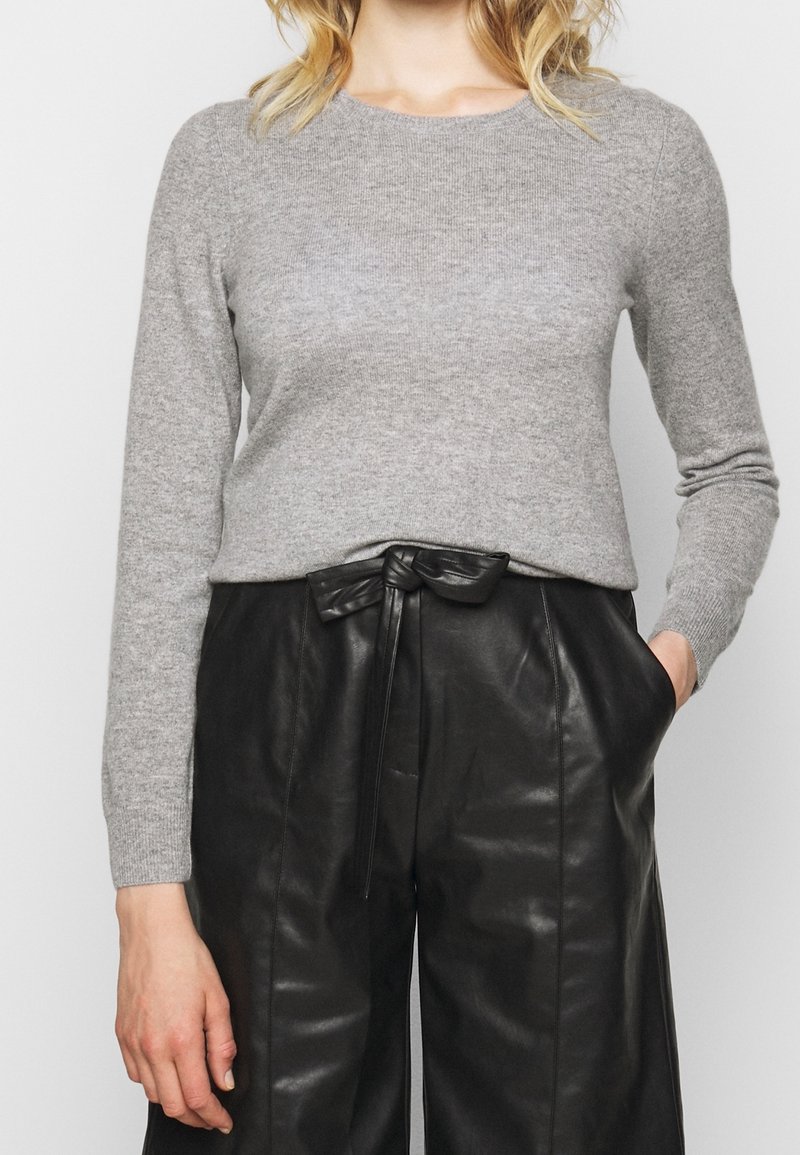 Femme portant un pull à manches longues gris clair rentré dans un pantalon en cuir noir taille haute avec une ceinture nouée et une main dans la poche.