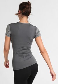 Mujer con cabello trenzado que lleva una camiseta deportiva de manga corta en tonos grises y leggings negros, de espaldas frente a un fondo liso.