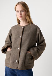 Brun hørfisket mønster jakke med rund hals, to forlommer og sølvtrykknapper. Har brede ærmer og beige trimdetaljer.