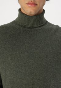 Pull à col roulé côtelé vert foncé en matière tricot douce, avec un design ajusté et un col haut structuré.