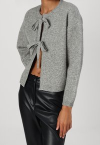Cardigan grigio lavorato a maglia con colletto rotondo, dotato di due legami frontali, spalle cadenti e polsini a coste. Indossato sopra pantaloni in pelle nera.