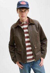 Giacca marrone con zip e colletto in corduroy, due tasche frontali, indossata sopra una polo a righe rossa, bianca e blu, abbinata a jeans blu.