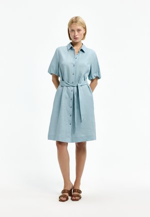 Woman standing with hands behind back wearing a light blue buttoned dress with a waist tie and brown heeled sandals.