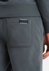 Graue Jogginghose mit einer Gesäßtasche mit rechteckiger Klappe und Markenlabel. Der Stoff scheint weich mit einer glatten Textur zu sein.