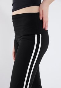 Leggings noirs avec une large ceinture noire et deux bandes verticales blanches sur les côtés extérieurs. Le tissu semble lisse et élastique.