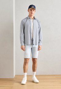 Chemise rayée, à manches longues, en bleu clair et blanc, portée sur un t-shirt blanc. Shorts en denim clair et baskets blanches, accompagnés d'une casquette bleu marine.