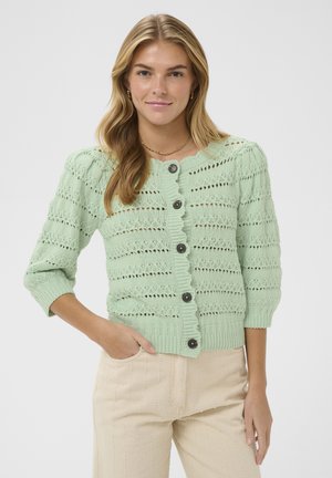 Lyshåret kvinde iført en lysegrøn strikket cardigan med sorte knapper og cremefarvede bukser, stående med den ene hånd i lommen og smilende blidt.