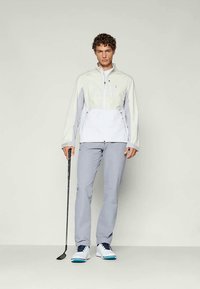 Veste de golf blanche et grise avec une fermeture éclair à l'avant, associée à un pantalon gris clair et des chaussures blanches avec des accents bleus. Tenant un club de golf.