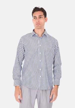 Giovane uomo che indossa una camicia a maniche lunghe con bottoni, a righe verticali blu navy e bianche, e pantaloni grigio chiaro, in piedi davanti a uno sfondo semplice.