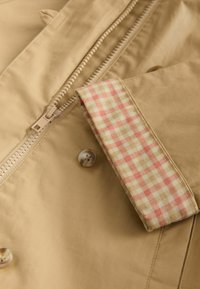 Bézs dzseki cipzárral, kerek gombbal és piros-bézs kockás szövetdísszel a galléron. Sima tapintású.