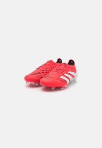 Red soccer cleats with a textured upper, featuring gray accents and three white stripes, designed for traction with visible studs.