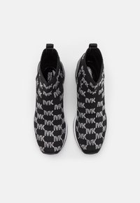 Ein Paar schwarze Slipper-Sneaker mit weißem "MK"-Logo-Muster und weißen Zwischensohlen, von oben auf einem hellen Hintergrund betrachtet.