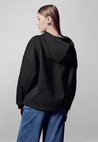 Felpa nera con cappuccio dal taglio rilassato, realizzata in materiale morbido. Abbinata a jeans a gamba larga blu per un look casual.