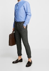 Lichtblauw overhemd met knopen, grijze getextureerde taps toelopende broek, zwarte leren loafers en een bruine leren tas.